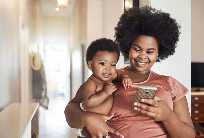 Young black mom holds her baby while smiling and looking at her mobile phone.