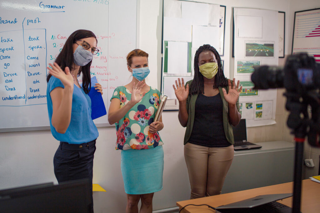Group of teachers with protective masks and standing near whiteboard wave to camera.