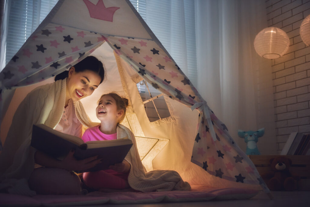 Mom and child daughter reading a book in tent under a soft glowing light.