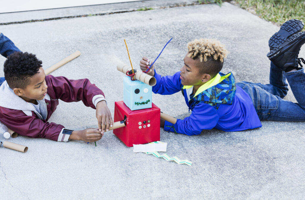 Two black middle school students play together outdoors on their driveway, building a robot out of cardboard