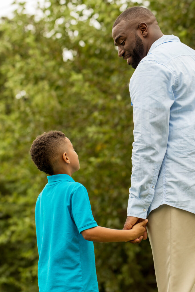 A father gazes down at his young son with a smile as they walk in the park.