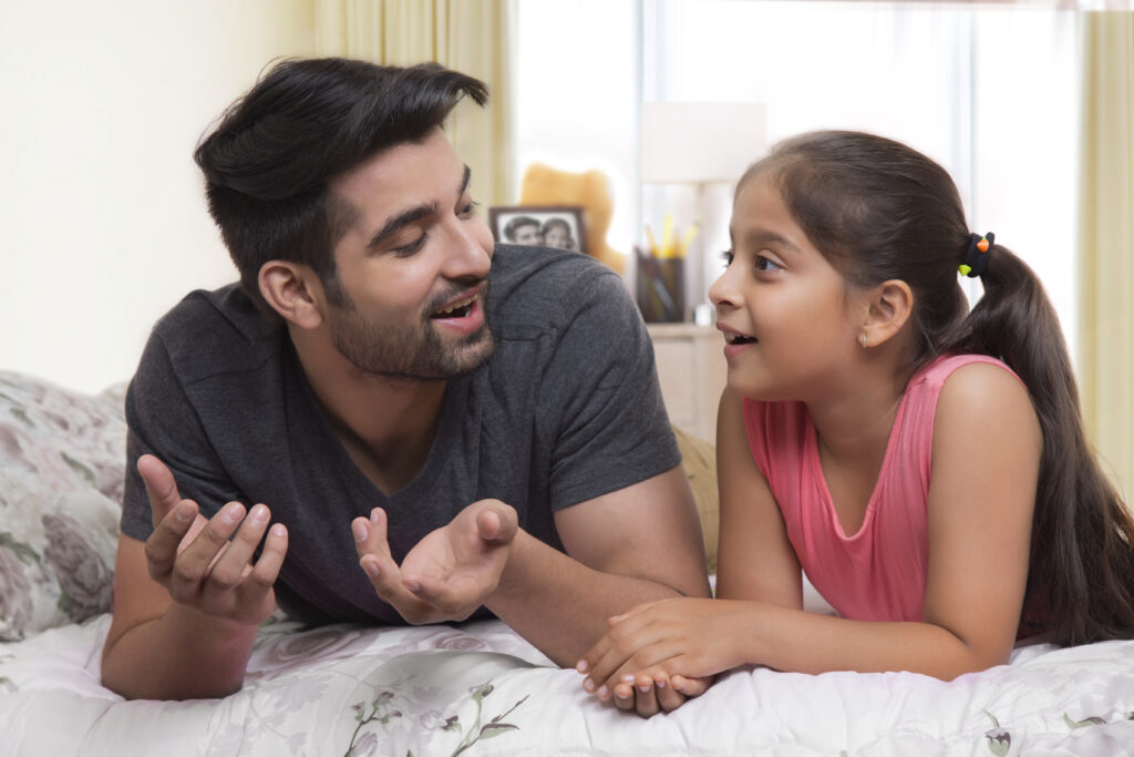 A father and daughter lie on her bed talking together and smiling. 