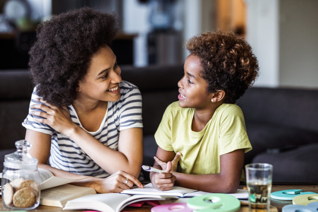 Young black girl talks with her mother as they work on homework at the kitchen table together. 