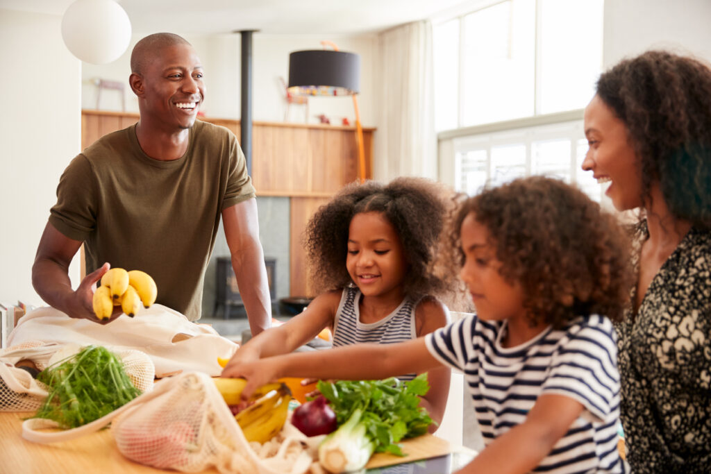 A smiling black family with a father, a mother, and two young daughters unpack grocery bags together in the kitchen. 