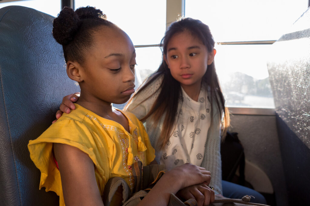 The young girl consoles her sad friend as they ride the bus to school.