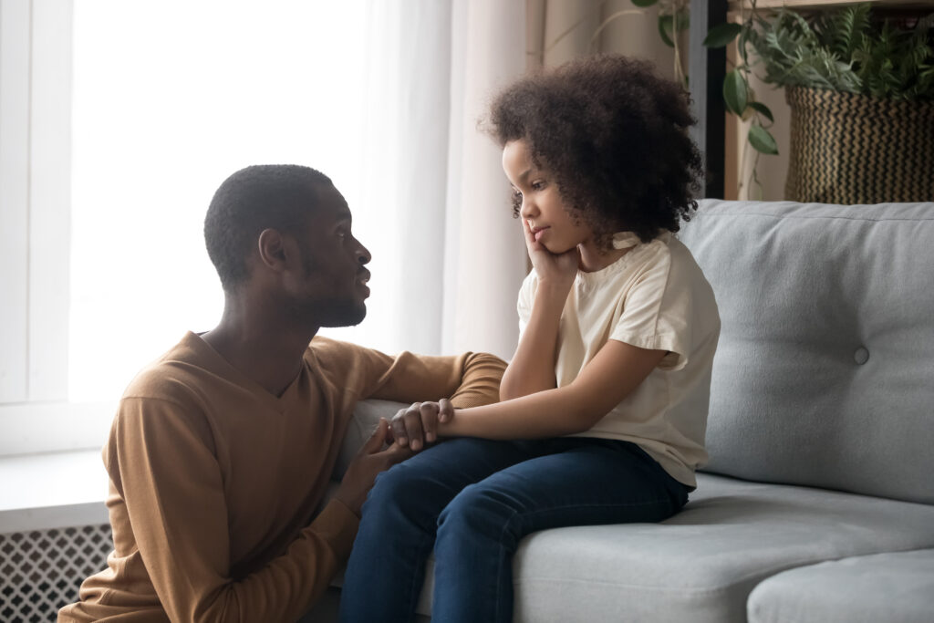 Black father comforts his young daughter, who looks sad and is sitting on the couch at home.