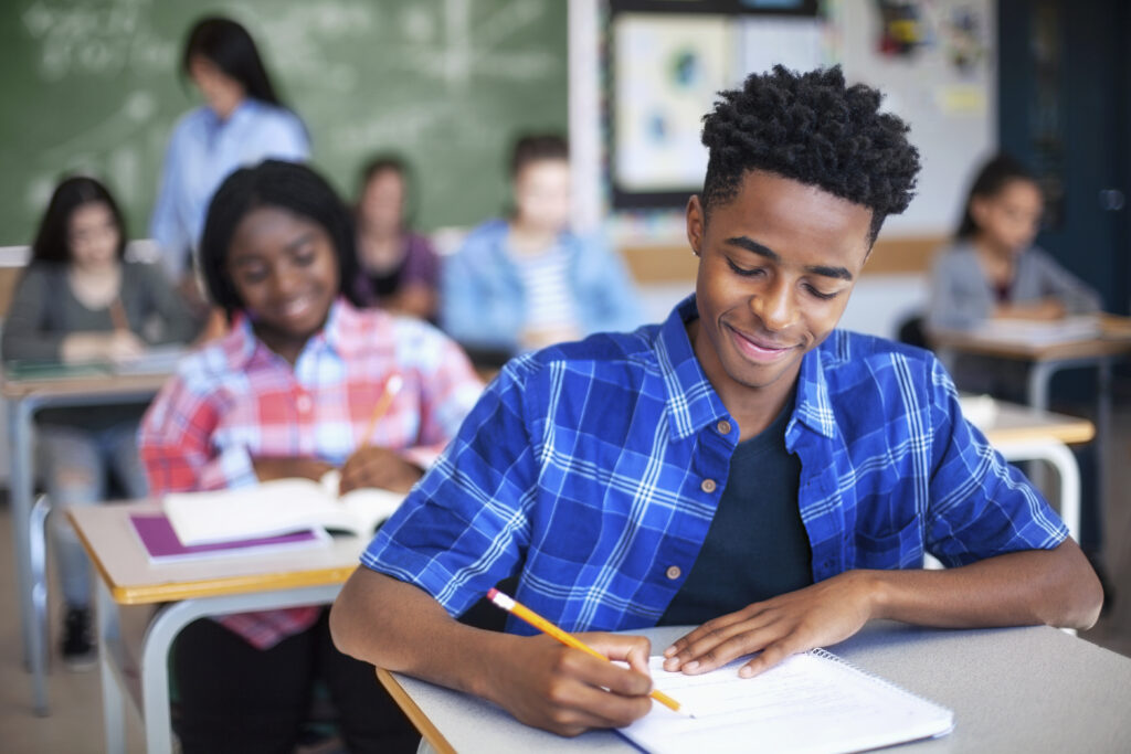 Smiling black teenager works on an assignment at his desk in school. 