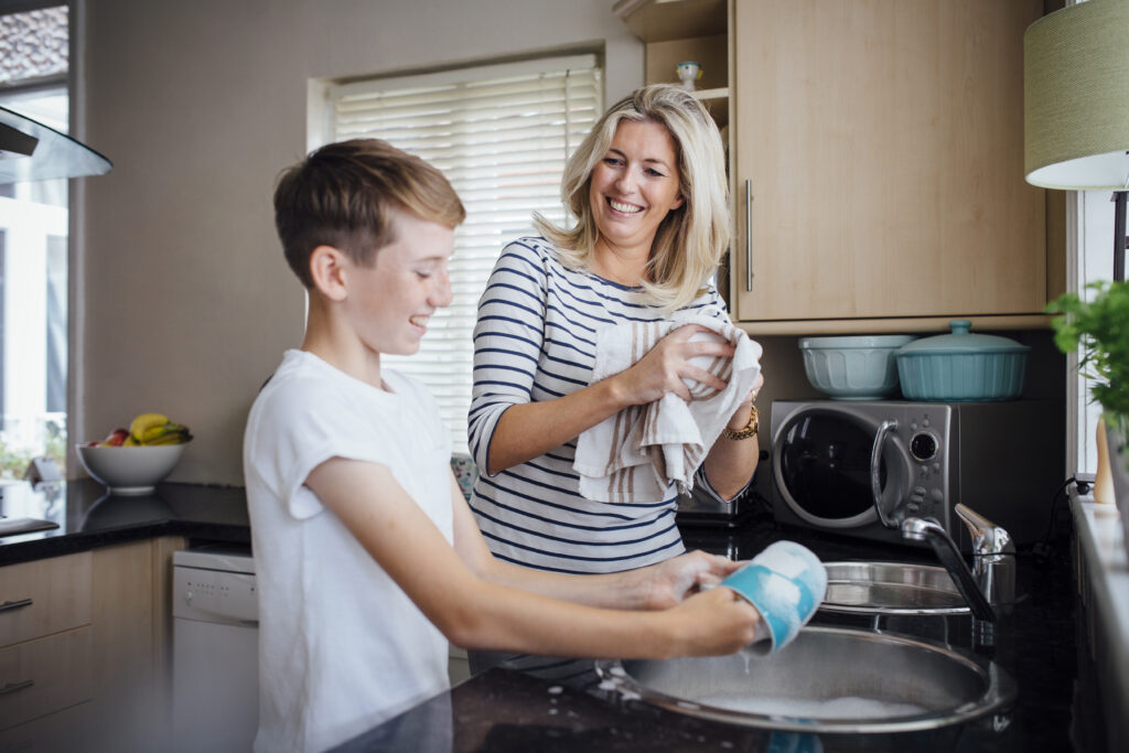 A Caucaisn mother and her son smile and wash dishes together in their kitchen.