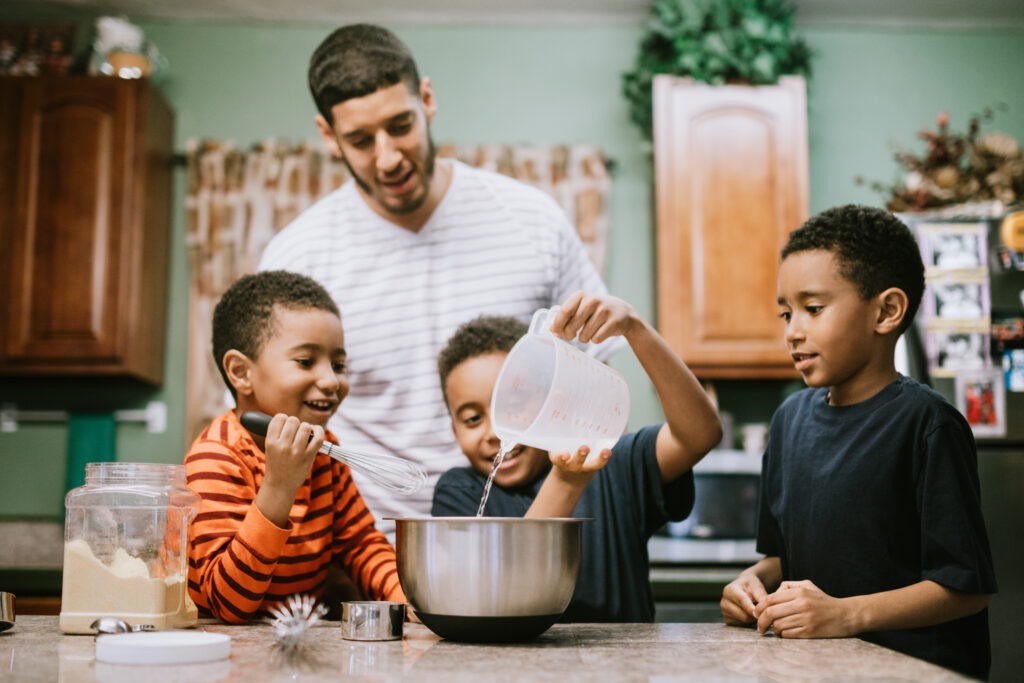 A dad and his three boys work together to make dinner at home, having fun spending family time together.  They mix and pour ingredients in to a bowl.