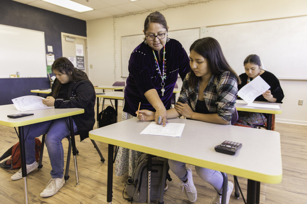 A Native American teacher helps a student at her desk with schoolwork. 