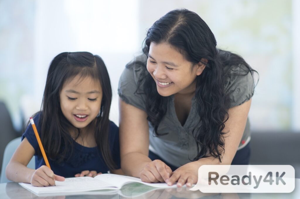 An Asian mother smiles while helping her young daughter with her homework.