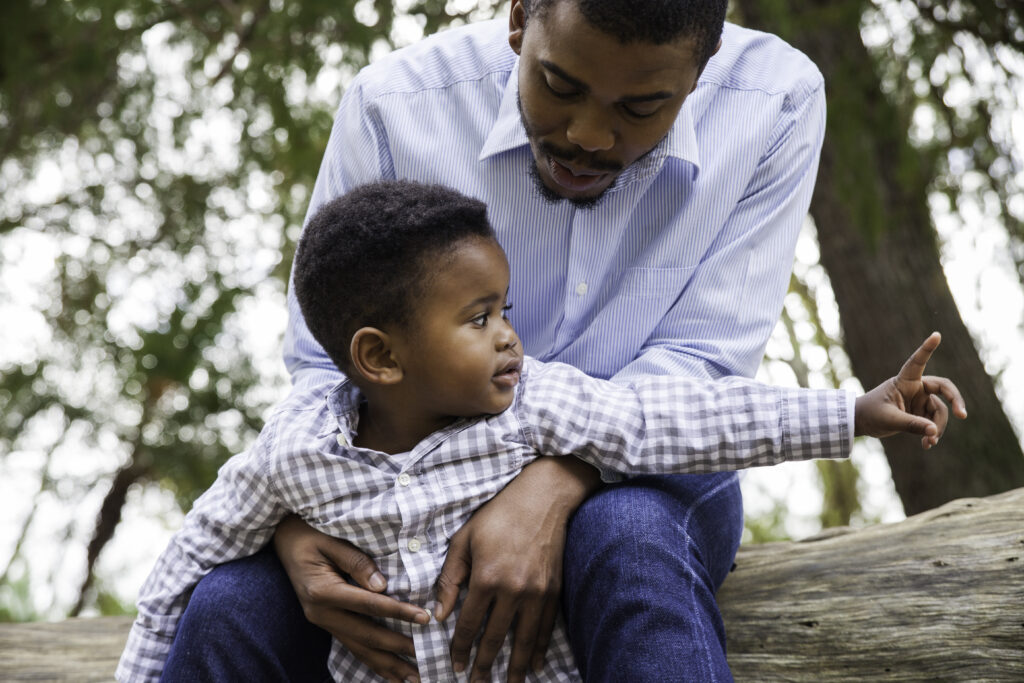 A black father sits with his toddler son as they explore a wooded park.