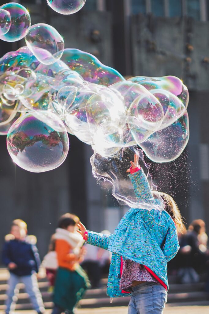 A young girl plays with giant bubbles in a city plaza surrounded by other children.