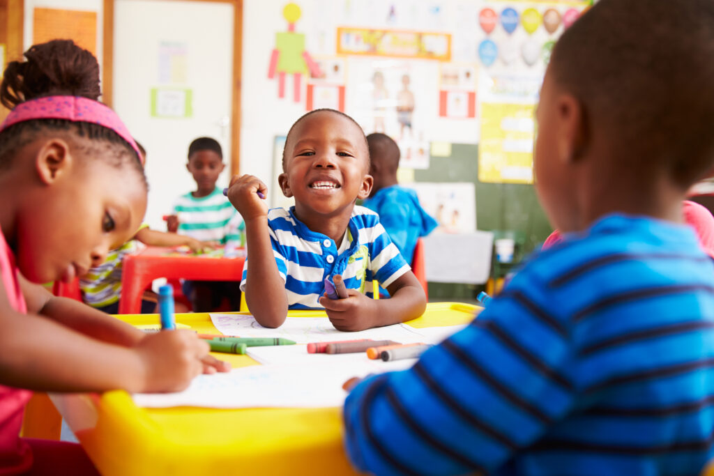 A black preschool boy smiles at the camera while working on an art project at school. 