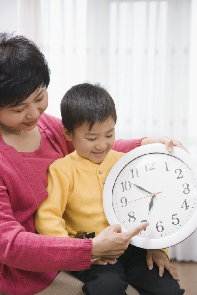 An Asian grandmother holds her preschool grandson in her lap as she shows him an analog clock.