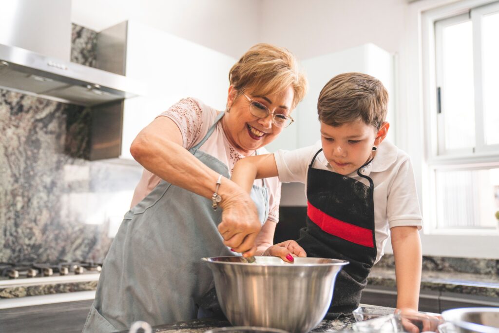 A mature Latin woman smiles as she helps her elementary school grandson stir ingredients into a mixing bowl in her kitchen.