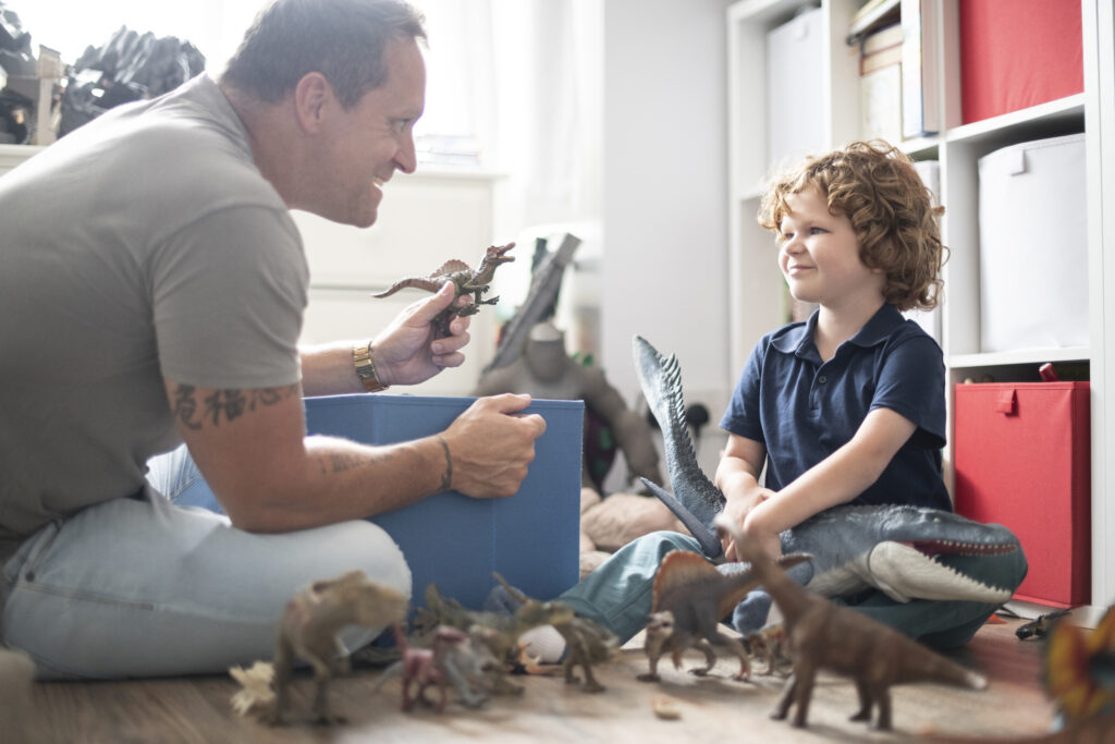 A mature father and his young son act out a story with toy dinosaurs on their living room floor. 