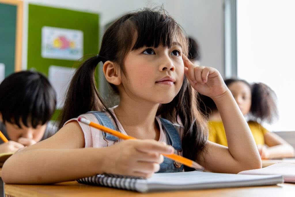 An Asian elementary school student holds her pencil over her notebook while thinking in class. 