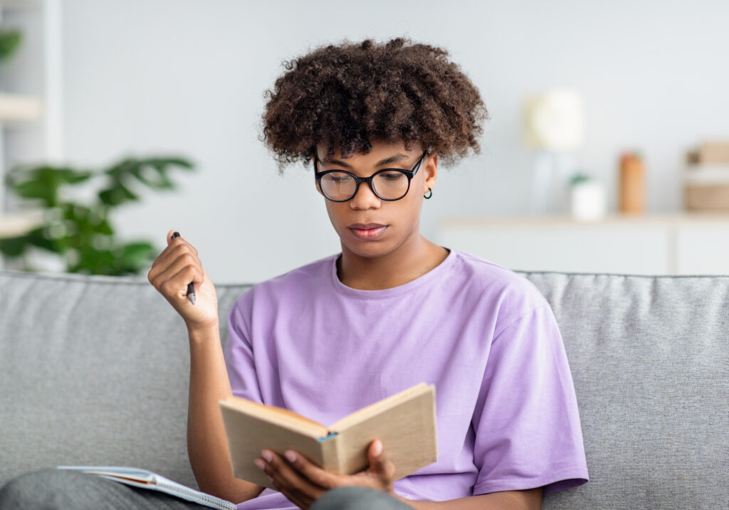 A black teenager holds a textbook while taking notes at home.