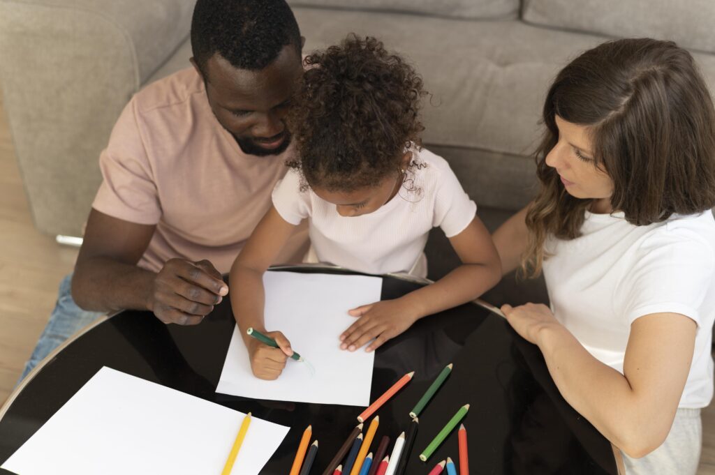 A multiethnic family gathers around a table at home. The father helps his young daughter with her drawing while the mother watches. 