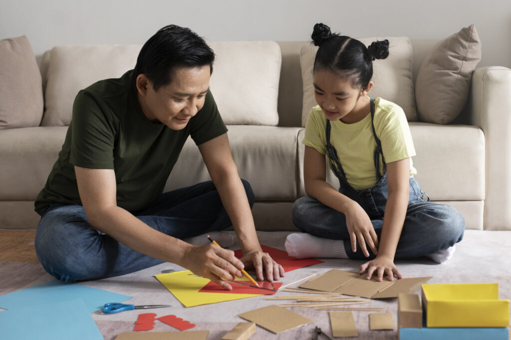 A mature Asian father helps his elementary school daughter with an art project at home.