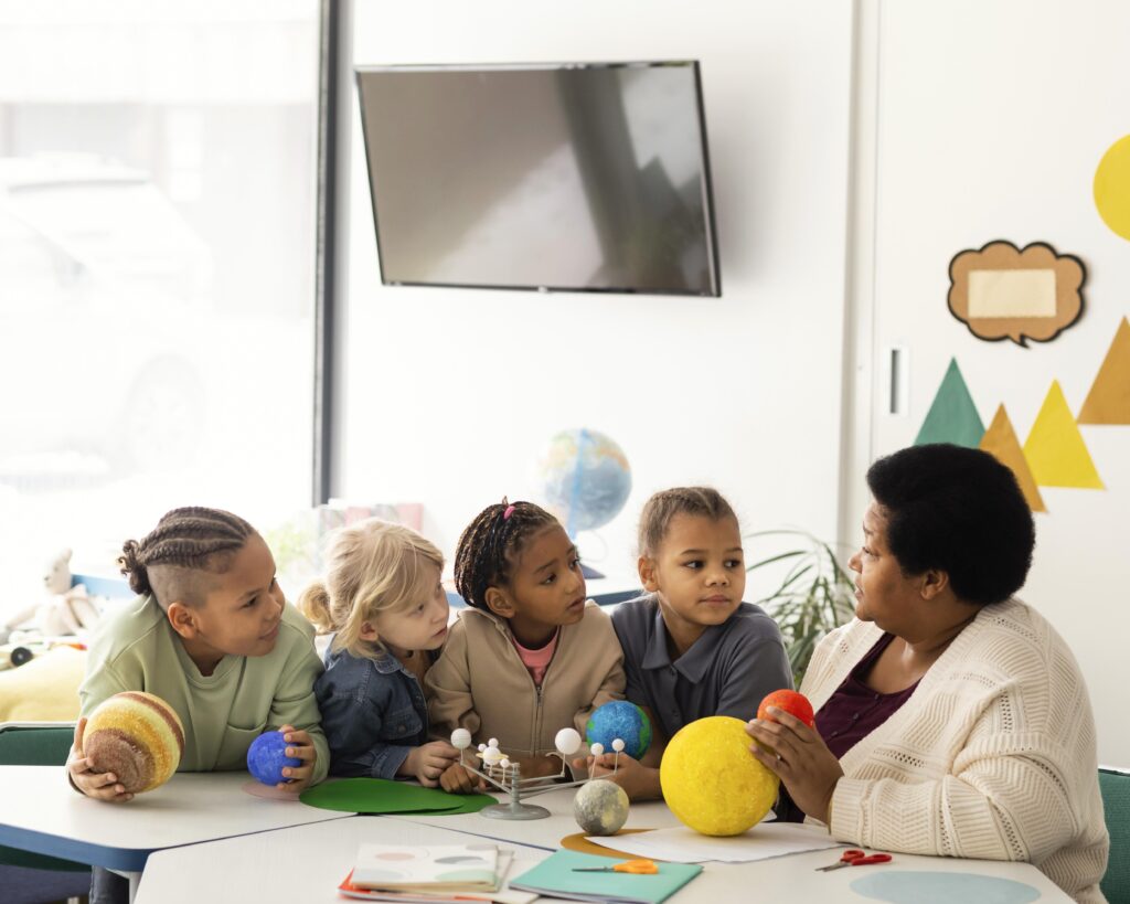 A black preschool teacher shows her diverse young students models of the planets in a classroom. 